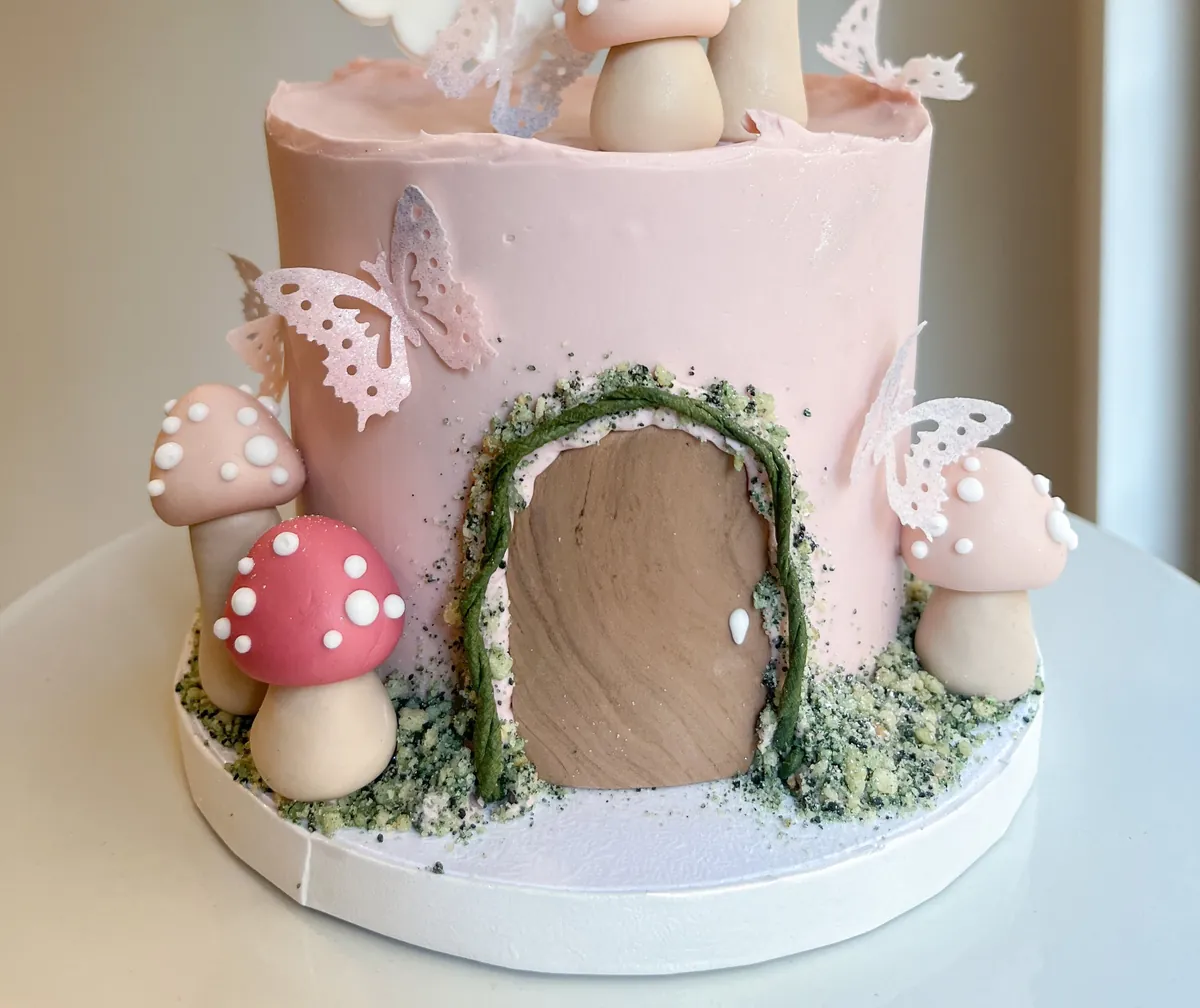 A pink cake with fondant toadstools, a carved fairy door and moss piping